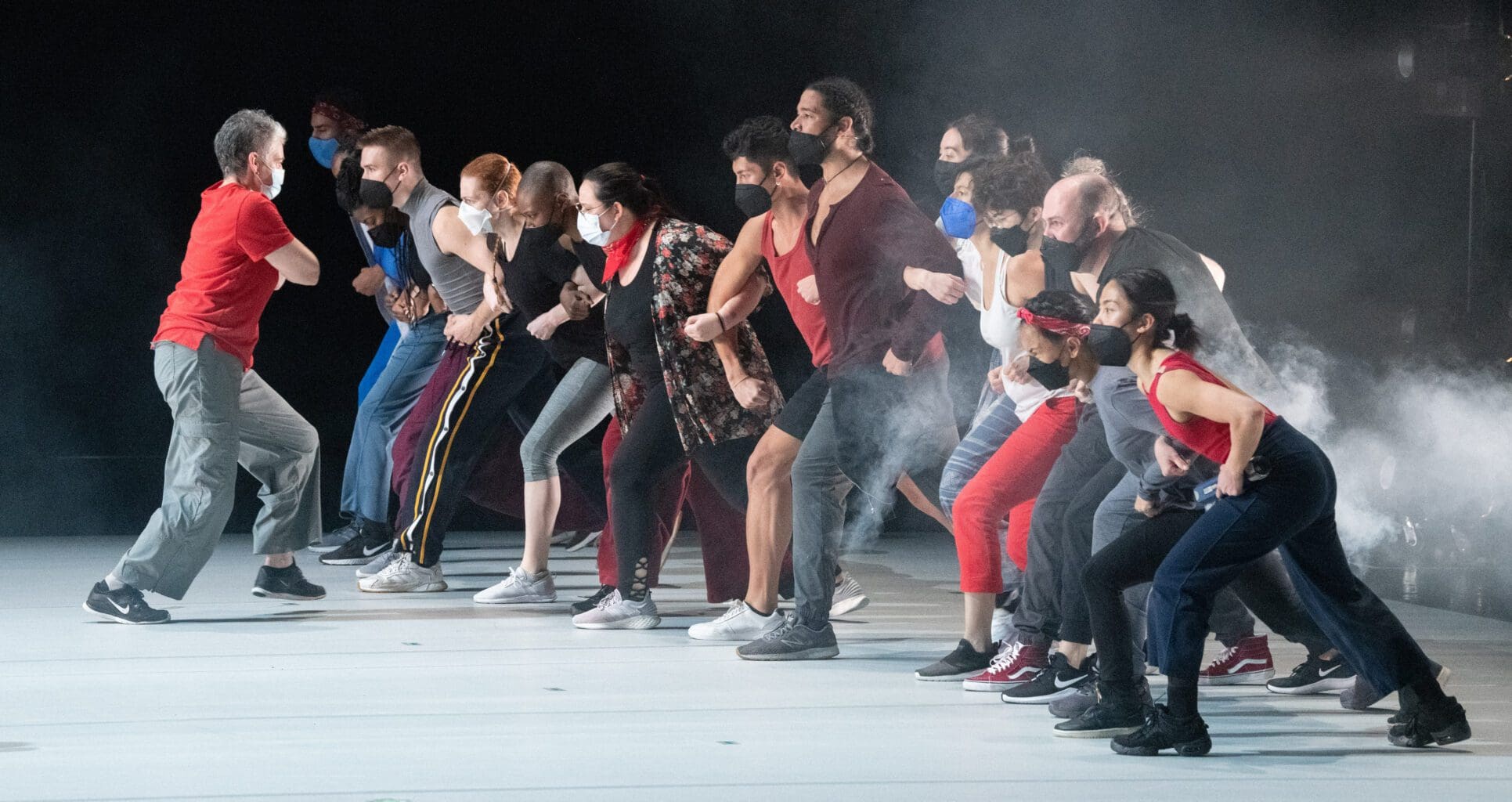 Photograph of group of performers on a white stage. They have their arms interlinked in a line, stepping forward in unison, as some white fog gathers behind them.