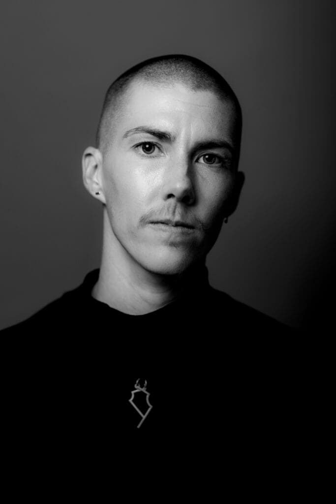 Black and white portrait of s.lumbert. They are wearing a black shirt with a small metal necklace.