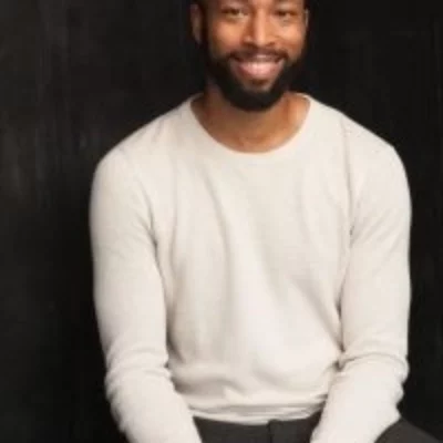 Medium headshot of Mark Hairston. They sit against a black background wearing a white sweater.