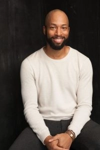 Medium headshot of Mark Hairston. They sit against a black background wearing a white sweater.