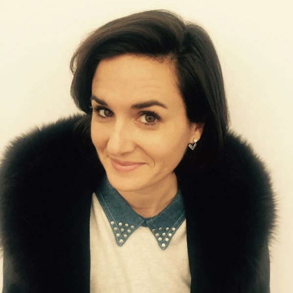 Color headshot photograph of Emily Manzo. They are smiling at the camera, wearing a white collared shirt and a black fur coat.