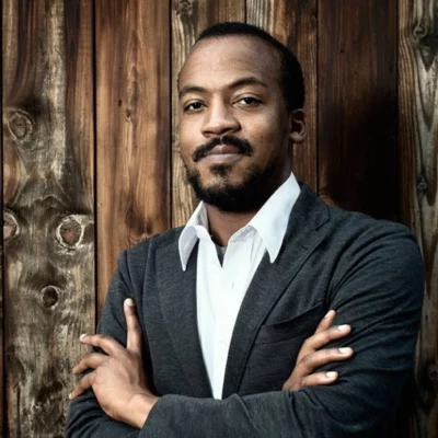 Color photo portrait of Matthew Gamble. They stand with arms crossed against a wooden wall, wearing a black suit and white collared shirt.