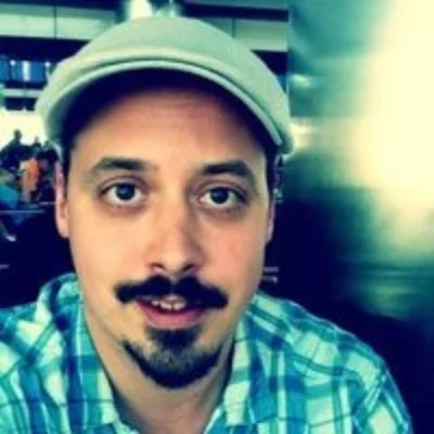 Selfie portrait of Sam Crawford. They are wearing a newscap and flannel shirt against a blurred background.