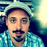 Selfie portrait of Sam Crawford. They are wearing a newscap and flannel shirt against a blurred background.