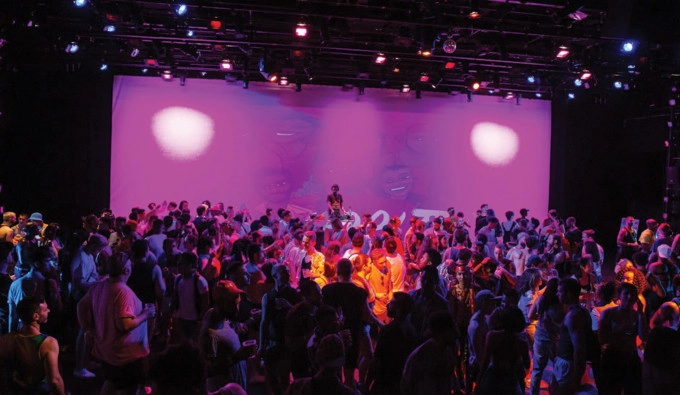 Large crowd in darkness on stage with a small group lit in orange, in front of a purple backdrop