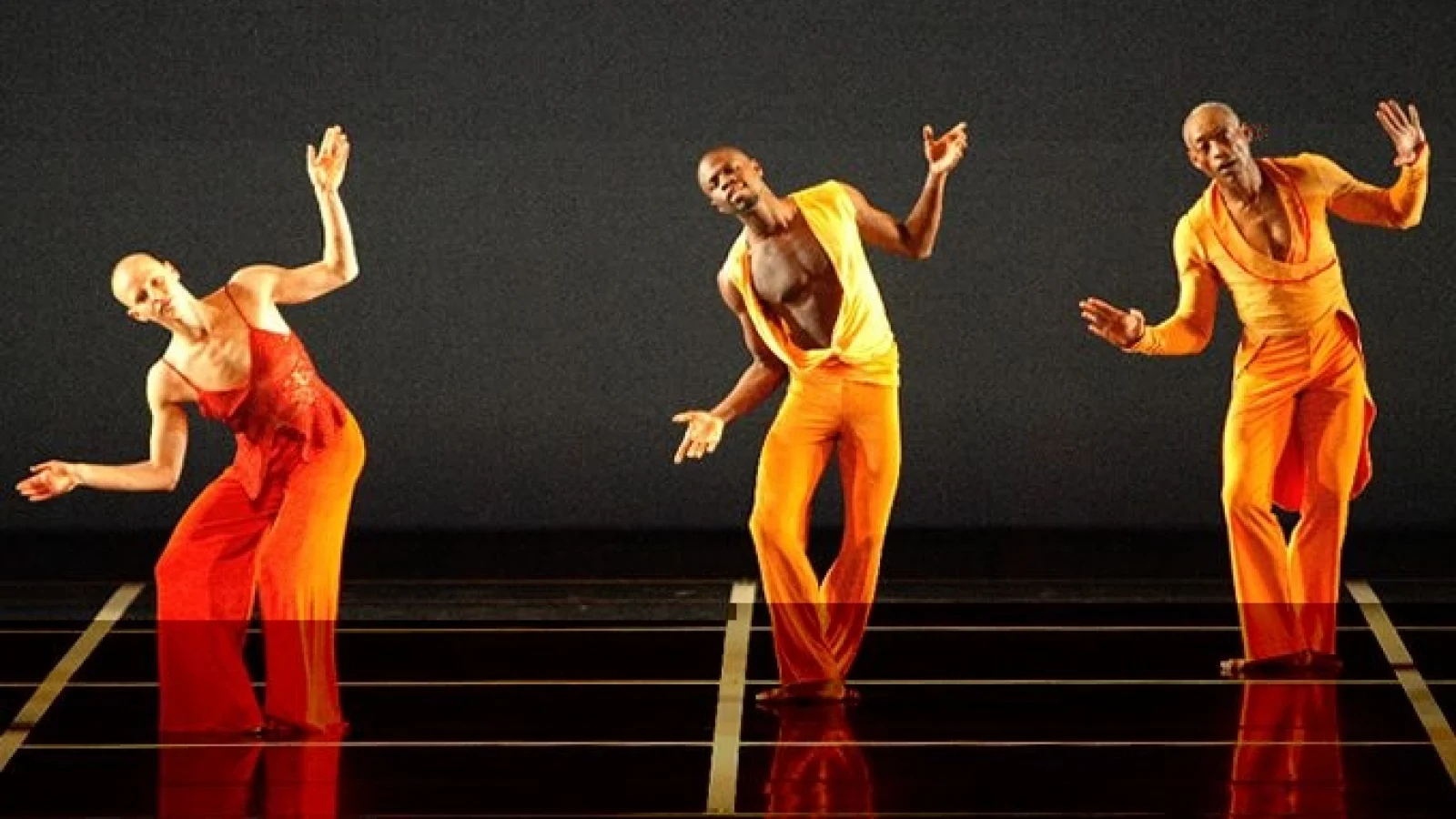 Three dancers in orange costumes tilt their bodies on a black stage