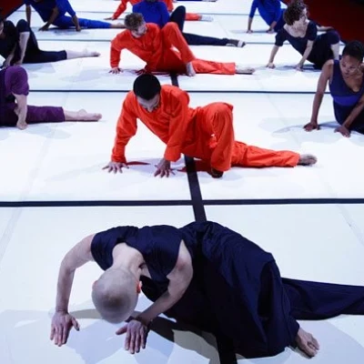 Three rows of dancers sit hunched over on a luminescent white stage