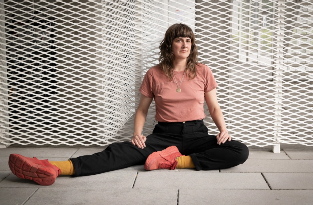 Medium shot of artist Faye Driscoll leaning against a white metal grate wall.
