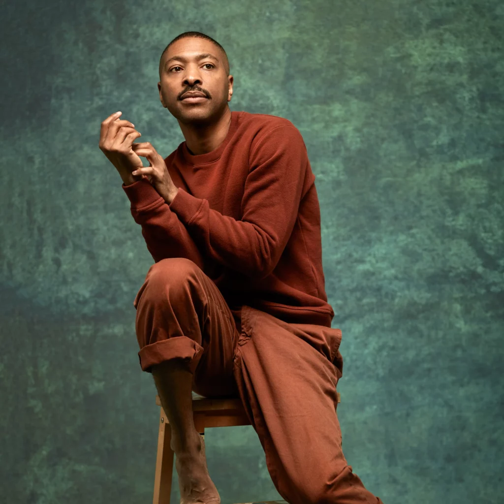 Studio portrait of Kyle Abraham. They are wearing an deep read sweater, umber pants, and are sitting pensively on a wooden stool against a textured green background.