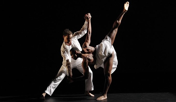 Two Figures Dressed in White Linen Dancing on the stage of a black box theater. One figure has dipped their body sideways while the other supports.