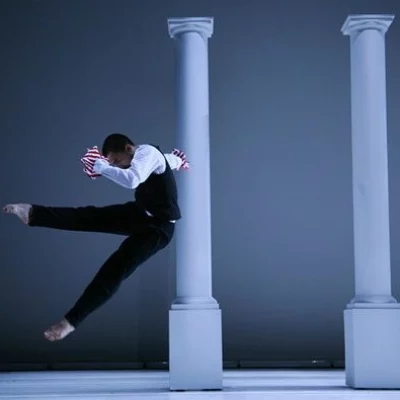 A dancer jumps and kicks their leg in front of a row of white pillars